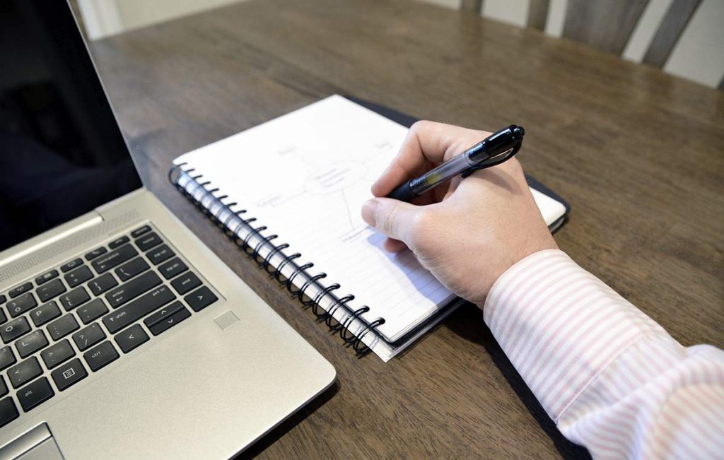 Brazo escribiendo en un cuaderno, sobre un escritorio con computador